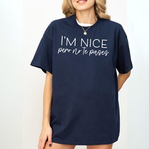 May include: A navy blue short-sleeved t-shirt featuring white text printed on the chest. The text reads I'M NICE in a bold sans-serif font, followed by the phrase pero no te pases in a smaller, cursive script font centered below it.