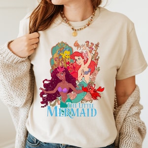 May include: A beige t-shirt featuring a vintage-style graphic of Ariel, Ursula, and Flounder from The Little Mermaid. The text "The Little Mermaid" is printed in blue below the image.