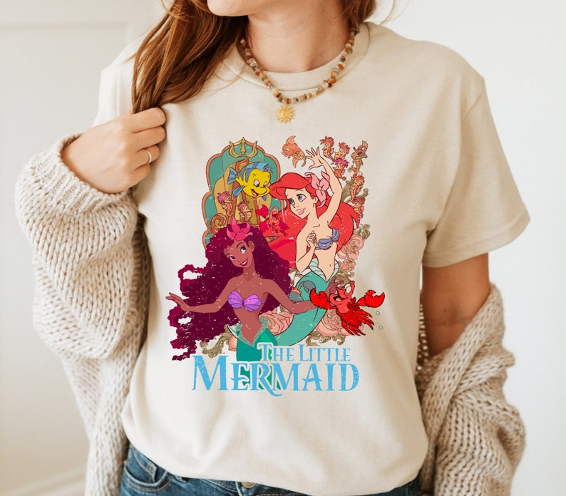 May include: A beige t-shirt featuring a vintage-style graphic of Ariel, Ursula, and Flounder from The Little Mermaid. The text "The Little Mermaid" is printed in blue below the image.