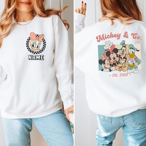 Op de afbeelding: Witte sweatshirt met een Daisy Duck-afbeelding, roze strik en de naam "NAME" in een zwart-wit geblokte cirkel. De achterkant toont een kleurrijk Mickey & Co.-ontwerp met Mickey, Minnie, Goofy, Donald en Daisy Duck, met "- Est. 1928 -" eronder.