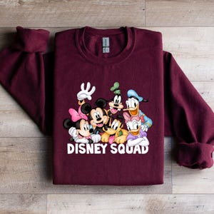 Puede incluir: Una sudadera burdeos con un gr&aacute;fico colorido de personajes de Disney, incluyendo Mickey Mouse, Minnie Mouse, Goofy, Donald Duck y Daisy Duck. Las palabras "DISNEY SQUAD" est&aacute;n impresas en blanco. La sudadera tiene mangas largas.