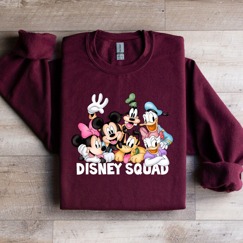 Puede incluir: Una sudadera burdeos con un gr&aacute;fico colorido de personajes de Disney, incluyendo Mickey Mouse, Minnie Mouse, Goofy, Donald Duck y Daisy Duck. Las palabras "DISNEY SQUAD" est&aacute;n impresas en blanco. La sudadera tiene mangas largas.