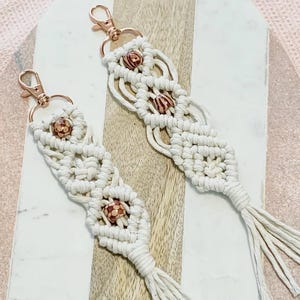 May include: Two macrame keychains with rose gold clasps. The keychains are off-white with decorative knots and small, round, brown beads. Each keychain has a tassel at the bottom.