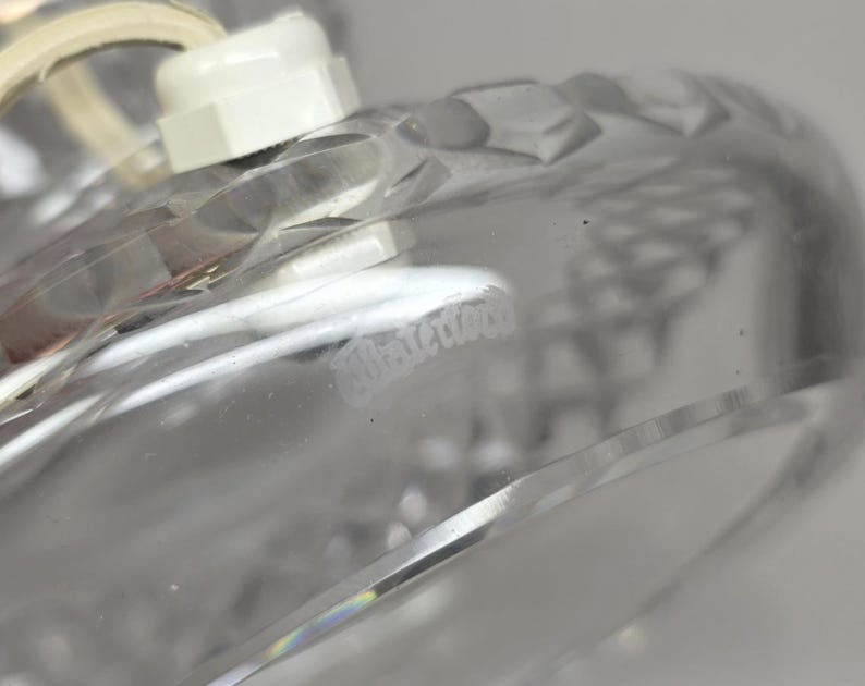 May include: Close-up of a clear glass object with a diamond-cut pattern. The object has a white plastic component attached to it, with a white cord. The word "Waterford" is faintly visible on the glass.