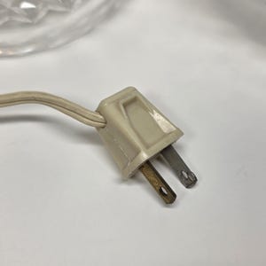 May include: Close-up of a beige electrical plug with two metal prongs. The plug is connected to a beige electrical cord. The background is white.