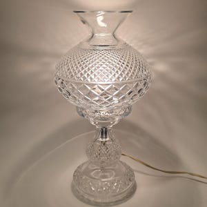 May include: A clear glass table lamp with a diamond-cut pattern on the globe-shaped shade. The lamp has a pedestal base and a flared top, with a visible power cord. The lamp is lit, casting a warm glow.