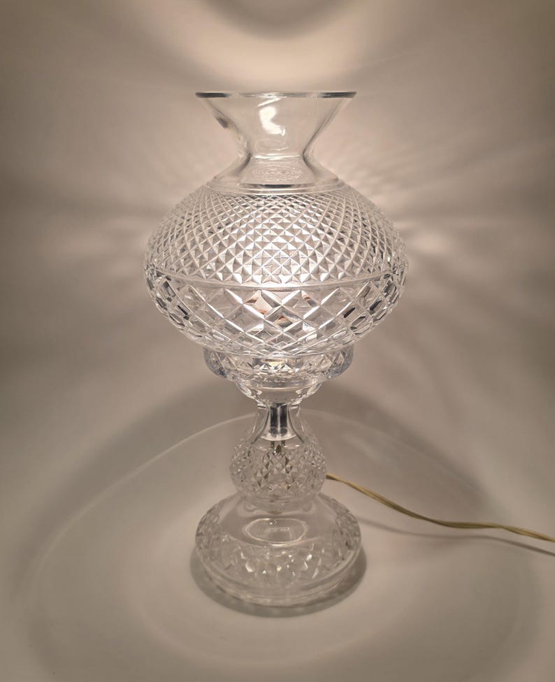 May include: A clear glass table lamp with a diamond-cut pattern on the globe-shaped shade. The lamp has a pedestal base and a flared top, with a visible power cord. The lamp is lit, casting a warm glow.