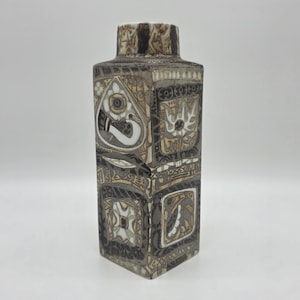 May include: A rectangular ceramic vase with a textured surface and a square base. The vase features a repeating pattern of geometric shapes in shades of brown, beige, and white. The top of the vase has a textured, patterned rim.
