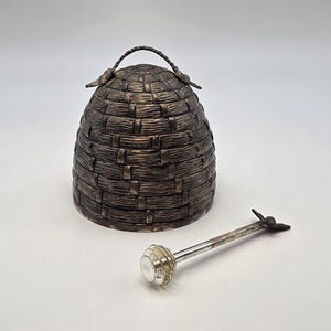May include: A silver-toned metal honey pot shaped like a traditional woven skep beehive, featuring a textured exterior and a small handle on top. Beside the pot lies a matching silver-toned honey dipper with a bee-shaped finial on the handle.