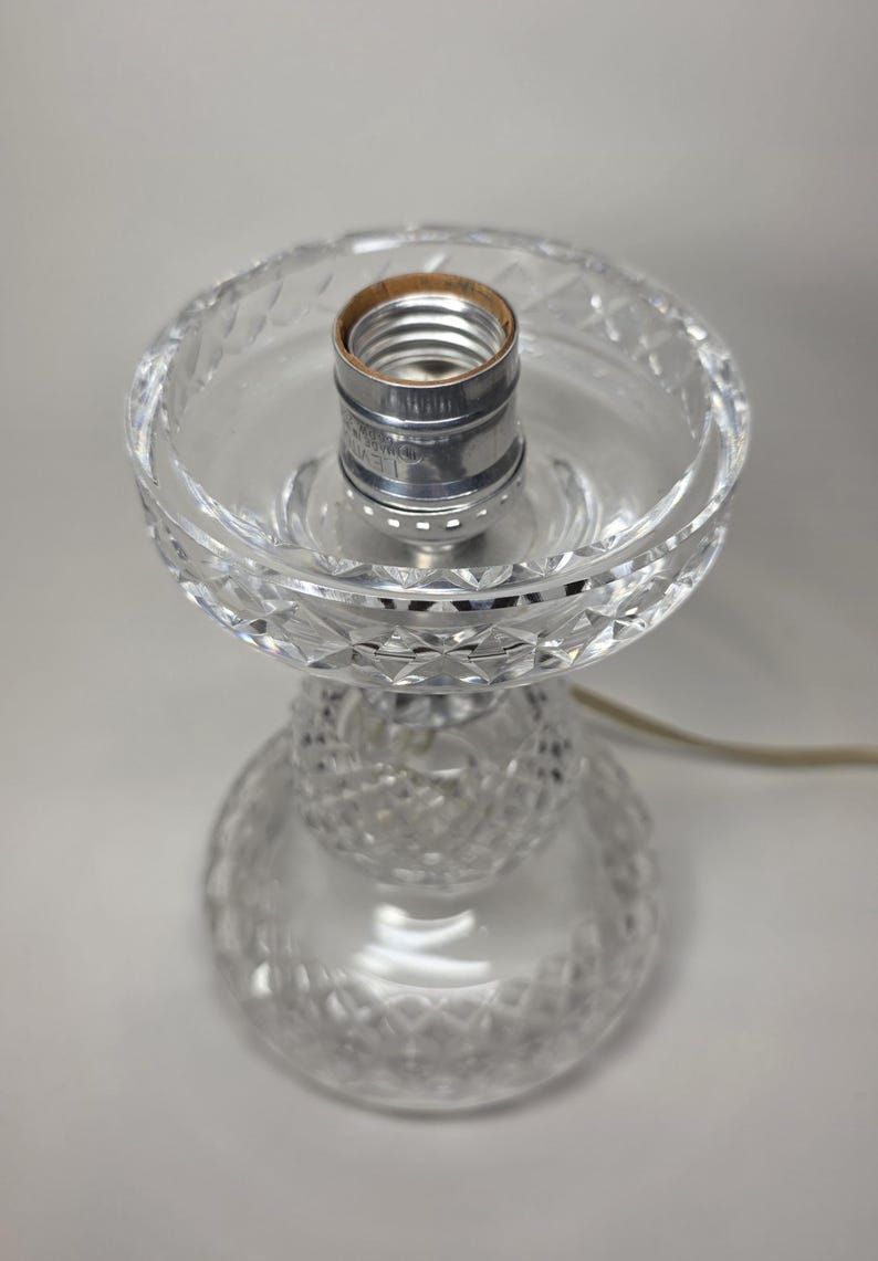 May include: A clear glass table lamp base with a decorative cut crystal design. The lamp base has a bulb socket and a power cord. The lamp base is approximately 8 inches tall.