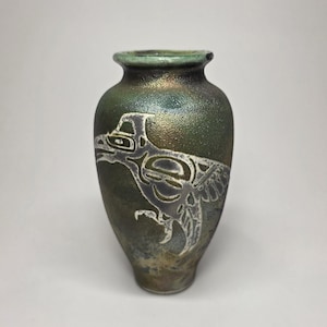 May include: A small, iridescent green vase with a detailed white design of a bird or mythical creature. The vase has a rounded body and a narrow neck, with a smooth, glossy finish. The design is etched into the surface.