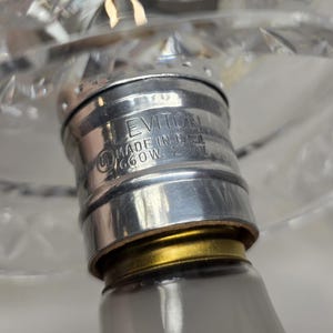 May include: Close-up of a lamp fixture with a silver-colored metal base. The base has the text "LEVITION MADE IN USA 660W 250V" etched into it. A clear, cut-glass lampshade is visible in the background.