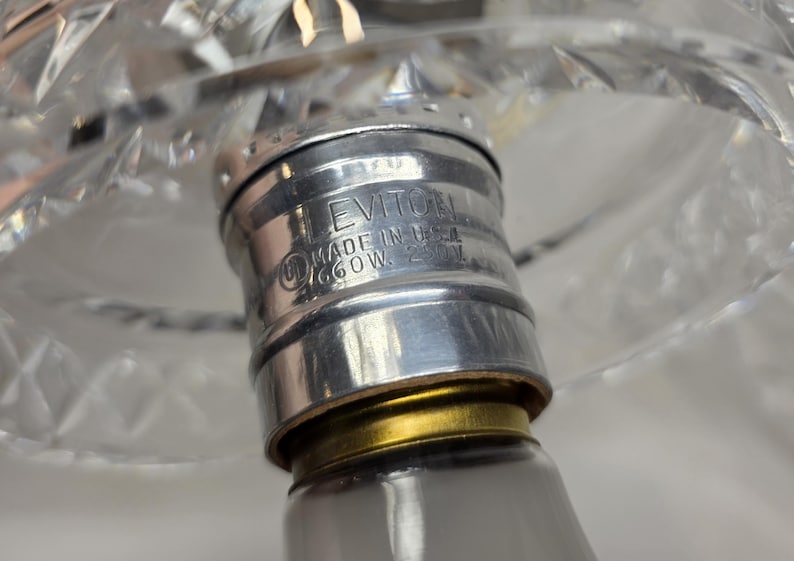 May include: Close-up of a lamp fixture with a silver-colored metal base. The base has the text "LEVITION MADE IN USA 660W 250V" etched into it. A clear, cut-glass lampshade is visible in the background.