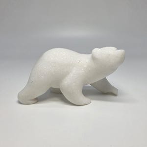 May include: A small, white, carved stone figurine of a bear in a walking pose. The sculpture features a smooth, polished surface with subtle crystalline flecks throughout the material, capturing the animal in a forward-leaning, dynamic stance.