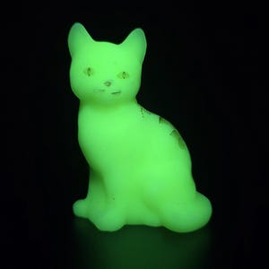 Peut inclure: Une petite figurine de chat assis en verre translucide qui brille d'une lumière vert néon éclatante. Le chat présente des oreilles pointues, des traits du visage définis et une forme de corps lisse et solide, lumineuse sur fond sombre.