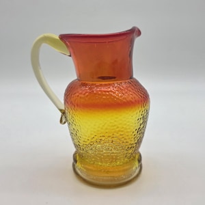 Peut inclure: Une cruche en verre vintage avec une surface texturée et un dégradé de couleurs. Le haut est rouge foncé, passant à l'orange puis au jaune. Elle a une anse crème et une base arrondie. Une pièce décorative pour servir des boissons.