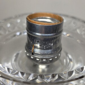 May include: Close-up of a clear glass lamp base with a cut crystal design. A silver metal lamp holder with a brown cardboard insert is in the center. The words "PRESS LAMP HOLDER" are printed on the metal.