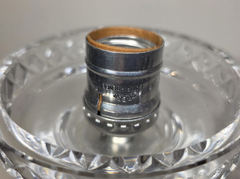 May include: Close-up of a clear glass lamp base with a cut crystal design. A silver metal lamp holder with a brown cardboard insert is in the center. The words "PRESS LAMP HOLDER" are printed on the metal.