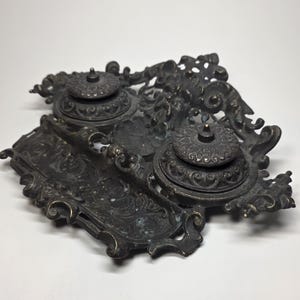 May include: An antique bronze inkwell set with two ink pots and a pen rest. The ink pots have ornate, domed lids. The set features intricate, swirling, and floral designs. The overall aesthetic is vintage and decorative.