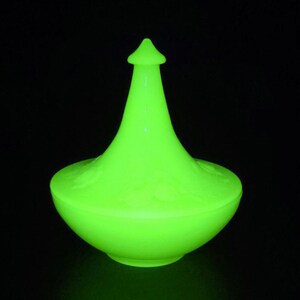 May include: A vibrant green glass jar with a lid, set against a black backdrop. The jar features a rounded base and a conical top, complete with a small finial. The surface of the jar has a subtle, embossed design.