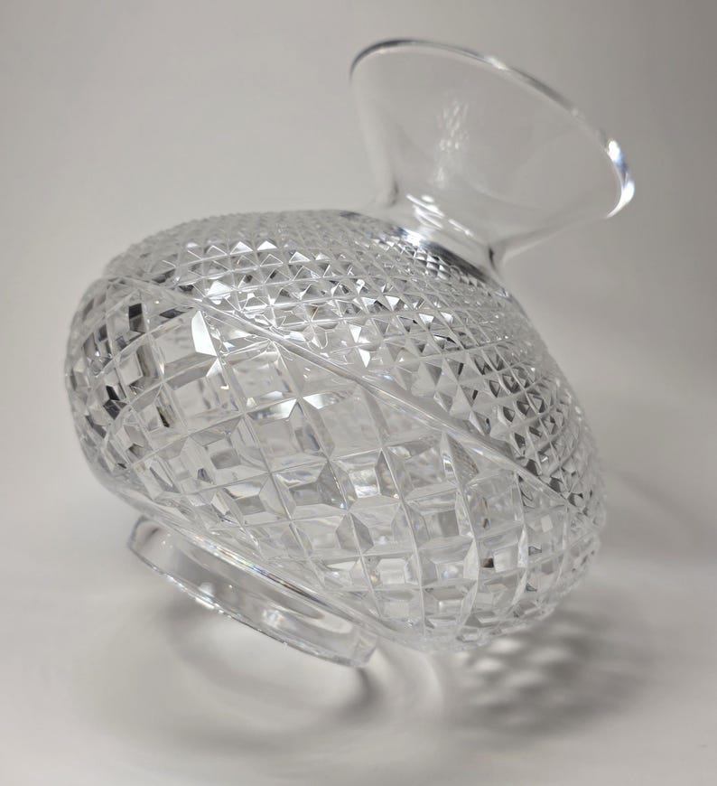 May include: Clear crystal pitcher with a diamond-cut pattern. The pitcher has a rounded body and a flared spout. The base is a circular shape. The pitcher is made of glass and is designed to hold liquids.