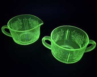 May include: Two vintage, glowing green glass serving pieces. One is a creamer with a spout, the other a sugar bowl with two handles. Both feature a floral pattern and are set against a black background.