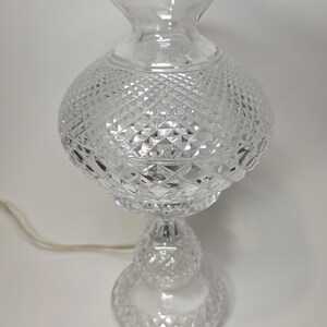 May include: Clear glass table lamp with a diamond-cut pattern. The lamp has a round base, a spherical midsection, and a flared top. A power cord extends from the base. The lamp is designed to provide ambient lighting.