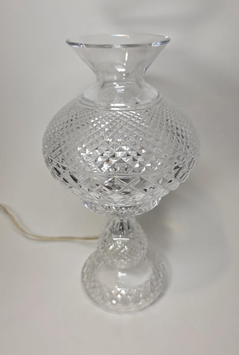 May include: Clear glass table lamp with a diamond-cut pattern. The lamp has a round base, a spherical midsection, and a flared top. A power cord extends from the base. The lamp is designed to provide ambient lighting.