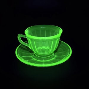Può includere: Un set di tazza e piattino verde brillante su sfondo nero. La tazza di vetro ha un manico e poggia su un piattino abbinato. Il set presenta un design strutturato attorno al bordo e alla base.