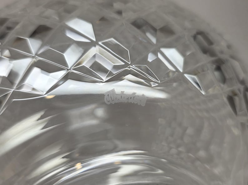 May include: Close-up of a clear glass object with a diamond-cut pattern. The glass has a reflective surface, and the word "Waterford" is etched into the glass. The object appears to be a decorative bowl or vase.
