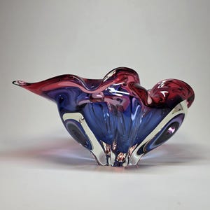 May include: A decorative glass bowl with a flowing, organic shape. The bowl features a blend of colours, including deep blue, vibrant pink, and a touch of white. The glass appears to be hand-blown, with a glossy finish.