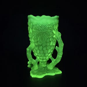 Peut inclure: Un vase en verre vert fluorescent avec un motif de grappe de raisin. Le vase présente une surface texturée, des anses en forme de branches et un bord festonné. La base est également texturée, sur fond noir.