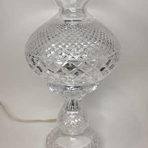 May include: A clear, cut-glass table lamp with a diamond pattern on the globe-shaped shade. The lamp has a flared top, a round base, and a decorative stem. The lamp is approximately 12 inches tall.