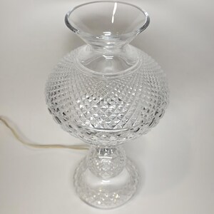 May include: A clear glass table lamp with a diamond-cut pattern on the globe-shaped body. The lamp has a flared top and a round base, with a power cord extending from the base. The lamp is approximately 12 inches tall.
