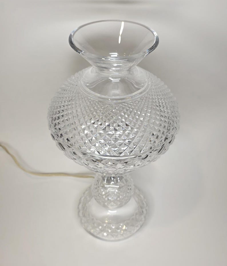 May include: A clear glass table lamp with a diamond-cut pattern on the globe-shaped body. The lamp has a flared top and a round base, with a power cord extending from the base. The lamp is approximately 12 inches tall.