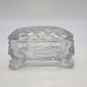 May include: Clear glass trinket box with a turtle design. The box has a lid and is supported by four small feet. The glass is intricately carved with a textured pattern, creating a decorative and elegant appearance.