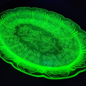 May include: An oval-shaped, translucent neon green glass platter featuring a central embossed design of a leafy branch with fruit, surrounded by a repeating floral pattern along the scalloped rim, glowing brightly against a dark background.