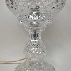 May include: A clear glass table lamp with a diamond-cut pattern. The lamp has a round base, a bulbous center, and a spherical shade. A power cord extends from the base. The lamp is a decorative lighting fixture.