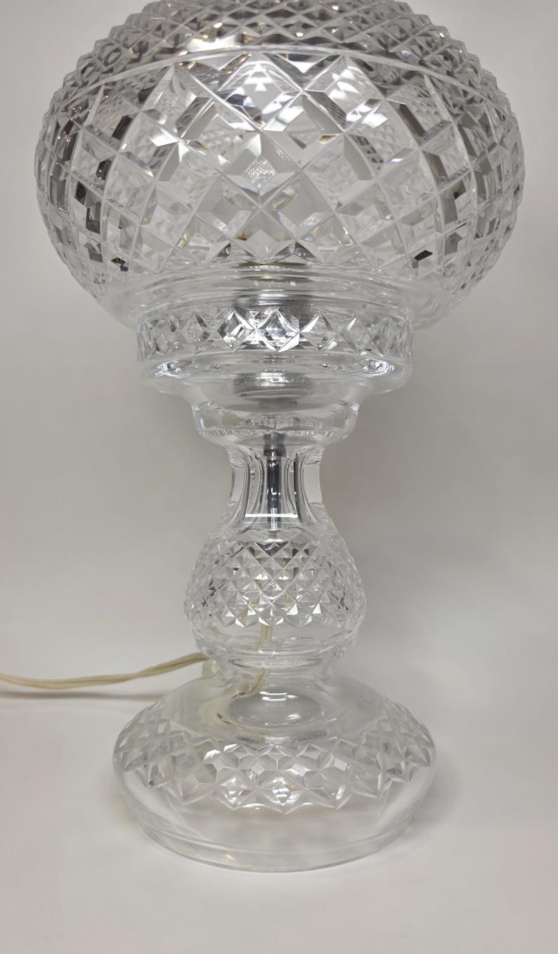 May include: A clear glass table lamp with a diamond-cut pattern. The lamp has a round base, a bulbous center, and a spherical shade. A power cord extends from the base. The lamp is a decorative lighting fixture.