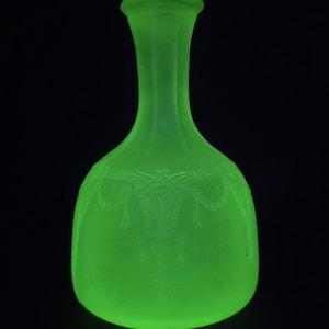 May include: A vibrant green glass vase, featuring a rounded base and a narrow neck. The vase has an etched decorative pattern around its body. Set against a black backdrop, the vase's bright colour and form are accentuated.