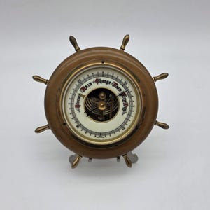 May include: A circular barometer shaped like a ship's wheel with a wooden frame and brass accents. The white dial features black markings, numbers, and the text Stormy, Rain, Change, Fair, and Very Dry, with visible internal mechanical components.