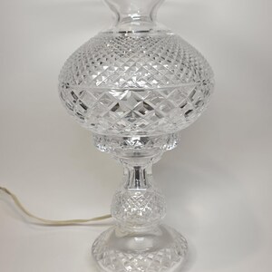 May include: Clear glass table lamp with a diamond-cut pattern. The lamp has a round base, a bulbous center, and a flared top. A power cord extends from the base. The lamp is a decorative lighting fixture.