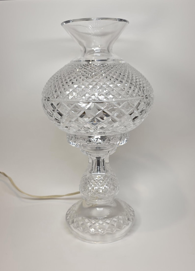 May include: Clear glass table lamp with a diamond-cut pattern. The lamp has a round base, a bulbous center, and a flared top. A power cord extends from the base. The lamp is a decorative lighting fixture.