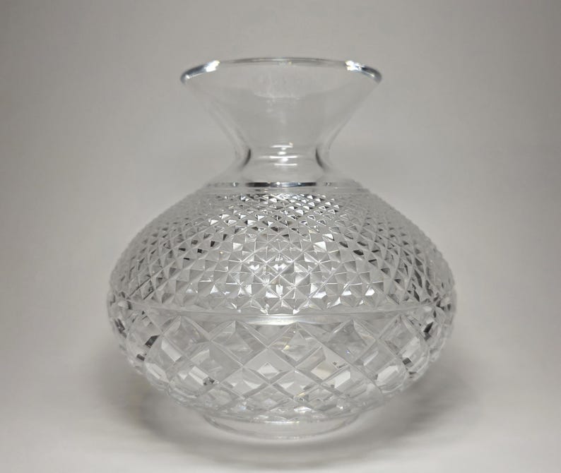 May include: A clear, cut-glass vase with a rounded body and a flared neck. The vase features a diamond-cut pattern across its surface, creating a textured and sparkling appearance. The vase is likely designed for decorative purposes or displaying flowers.