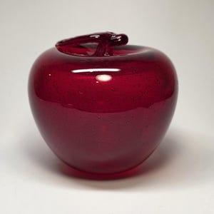 May include: A decorative, ruby-red glass apple. The apple has a smooth, glossy surface with a stem. The glass has small air bubbles throughout. This handcrafted piece is a unique home decor item.