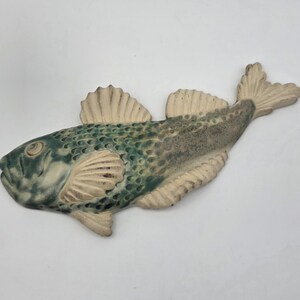 Peut inclure: Un ornement mural en céramique en forme de poisson avec un motif d'écailles texturé dans des tons de bleu sarcelle profond et de vert sourd, contrasté par des nageoires et une queue blanc cassé. La tête détaillée présente un œil et une bouche.