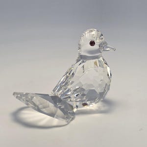 May include: A clear faceted crystal bird figurine featuring a pointed beak and a single small dark purple circular eye. The bird is depicted in a seated position with its wings folded against its body and a wide, flat tail extending outward.