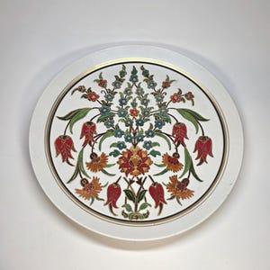 May include: A white, round decorative plate with a gold rim and a detailed floral design. The design features red, orange, blue, and green flowers and leaves. The plate has a classic, ornate style.