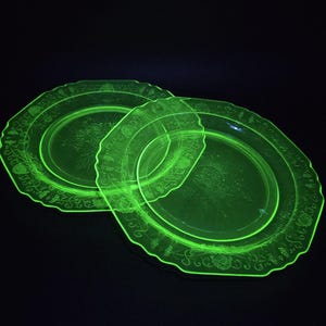 May include: Two round, translucent neon green glass plates featuring a scalloped edge and an embossed floral pattern along the rim. The plates glow with a bright, vivid light against a dark background, highlighting the intricate details.