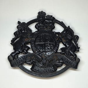 Peut inclure: Un emblème en fonte noire représentant des armoiries royales. L'emblème comprend une couronne, un écu divisé en quadrants et la devise "DIEU ET MON DROIT". Deux animaux flanquent l'écu.